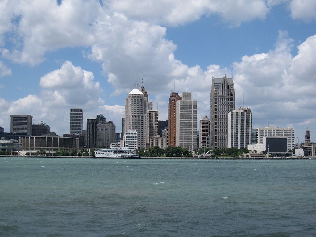 Downtown Detroit skyline seen across the Detroit River under a partly cloudy sky, with a white riverboat docked