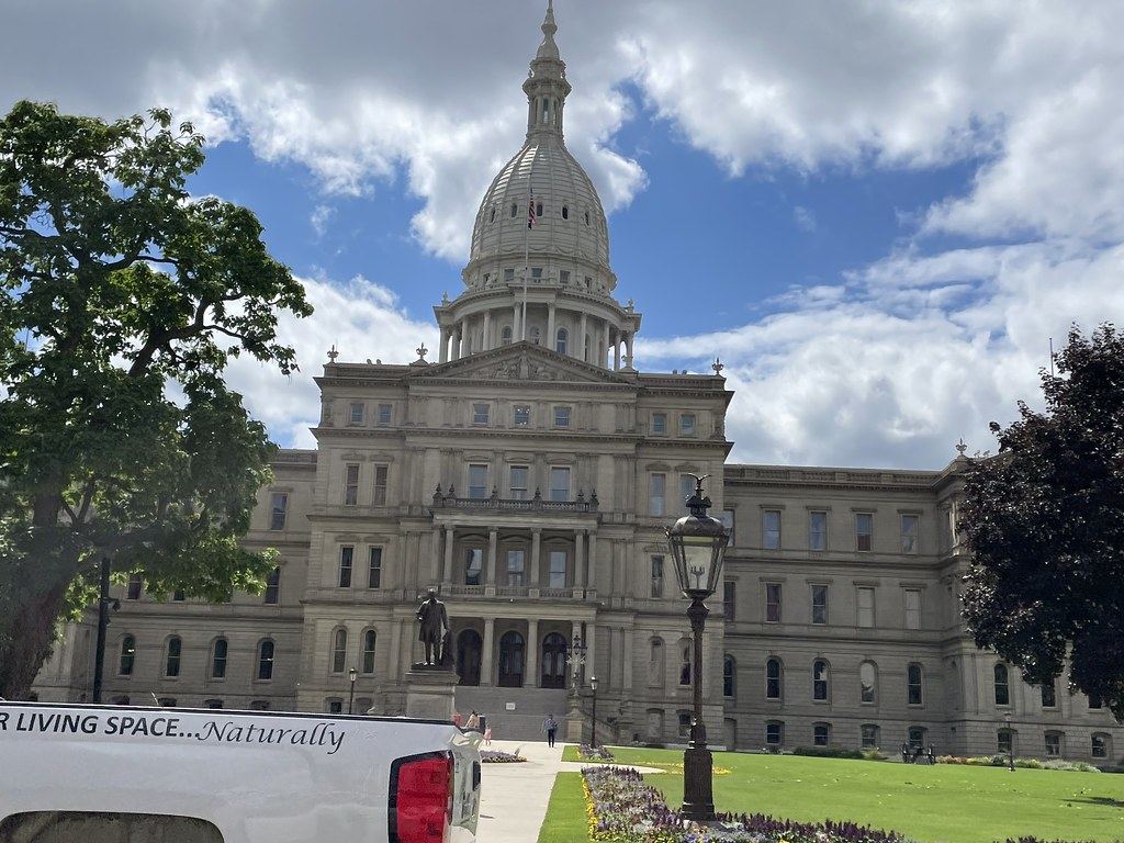 The domed Michigan State Capitol building with a statue, lamppost and flower beds on the lawn under a partly cloudy sky