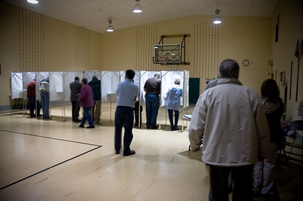 People stand at white-curtained voting booths in a school gymnasium while others wait in line