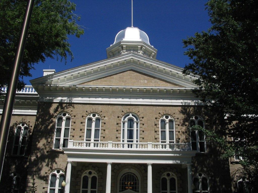 Sandstone capitol building with arched windows, white columns and a silver dome under a blue sky, framed by trees