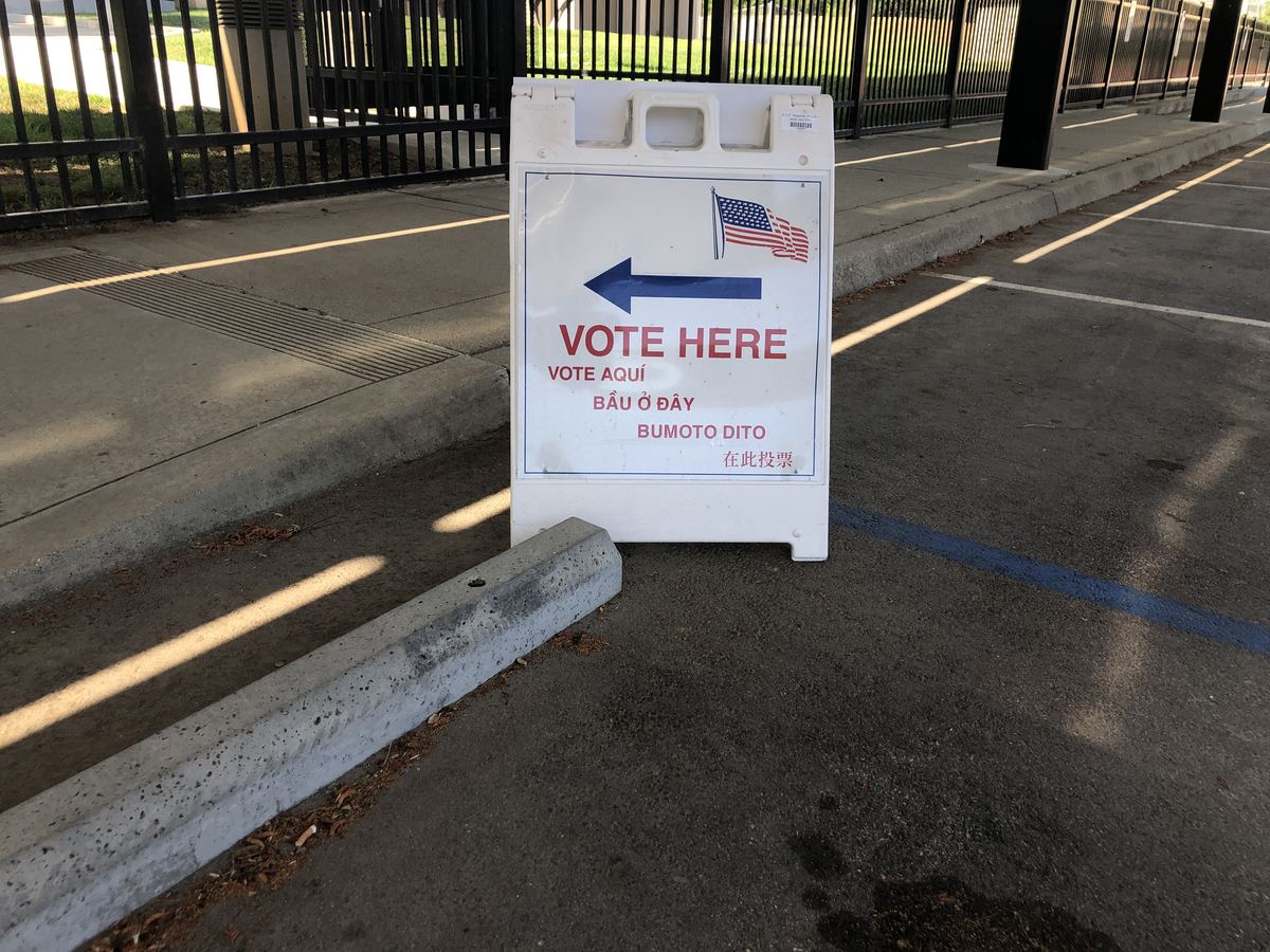 A multilingual 'Vote Here' sandwich board sign with an arrow and U.S. flag stands in a parking lot next to a fence