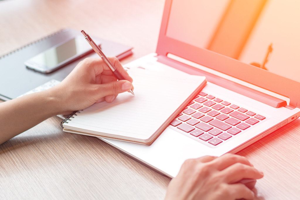 Hands writing in a spiral notebook resting on an open white laptop on a wooden desk beside a smartphone