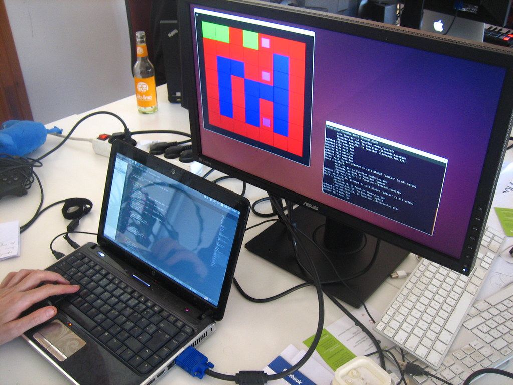 A laptop displaying code beside a monitor showing a colorful grid and a terminal window on a cluttered desk
