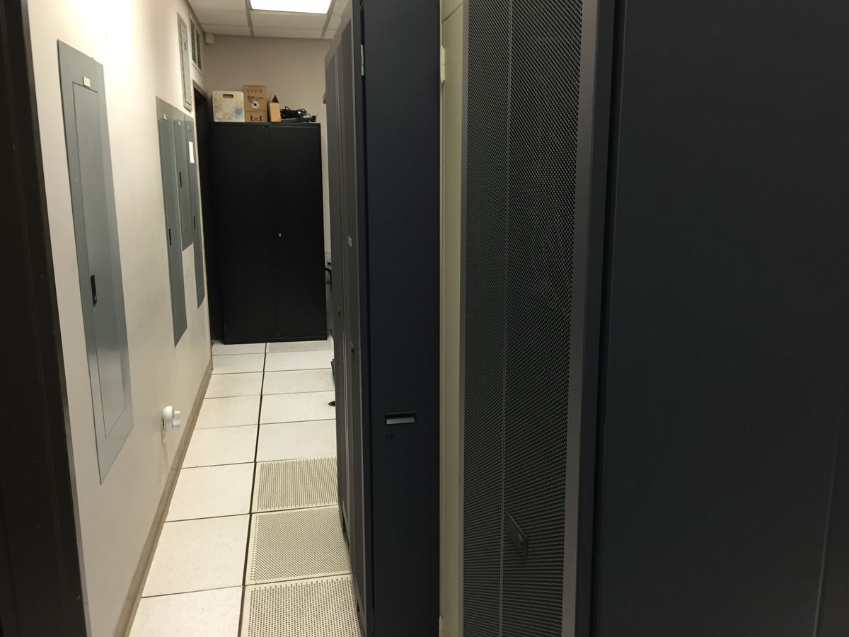 Narrow server room with dark rack cabinets, wall-mounted electrical panels and perforated raised floor tiles