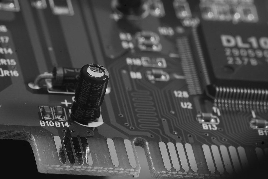 Black-and-white close-up of a circuit board with a capacitor, a chip and gold edge connector contacts