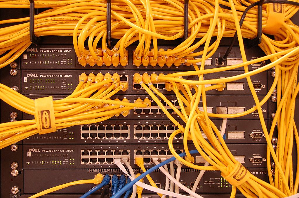 Rack of network switches with dozens of yellow and blue Ethernet cables plugged in and bundled with straps