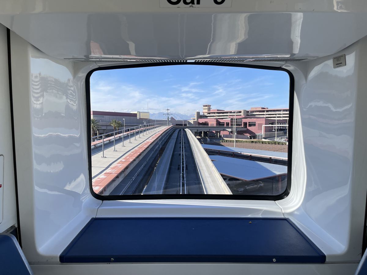 View from the front window of an elevated airport tram, with guideway tracks leading toward terminal buildings and mountains