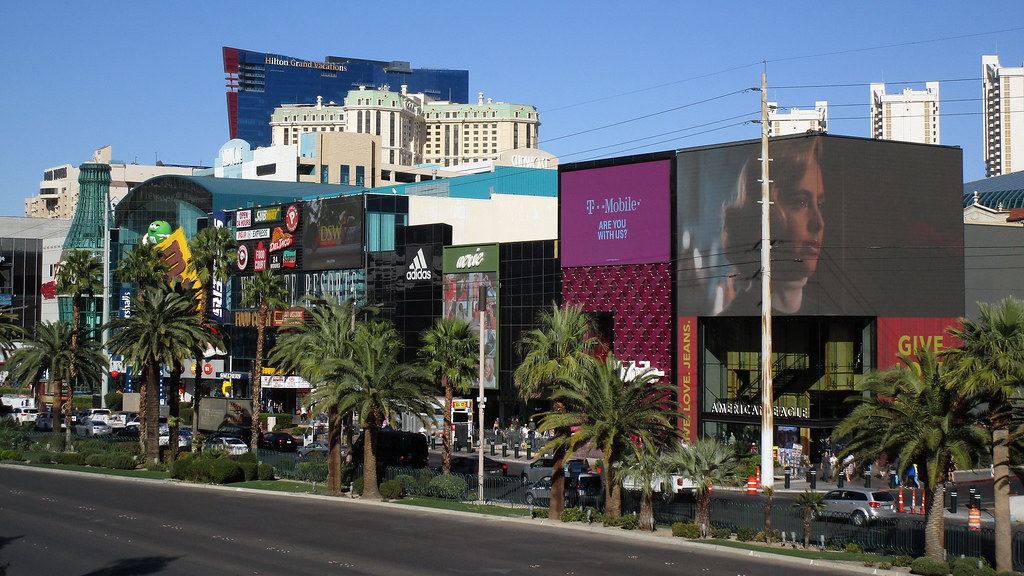 Las Vegas Boulevard lined with palm trees, shops, large billboards and hotel towers under a clear blue sky