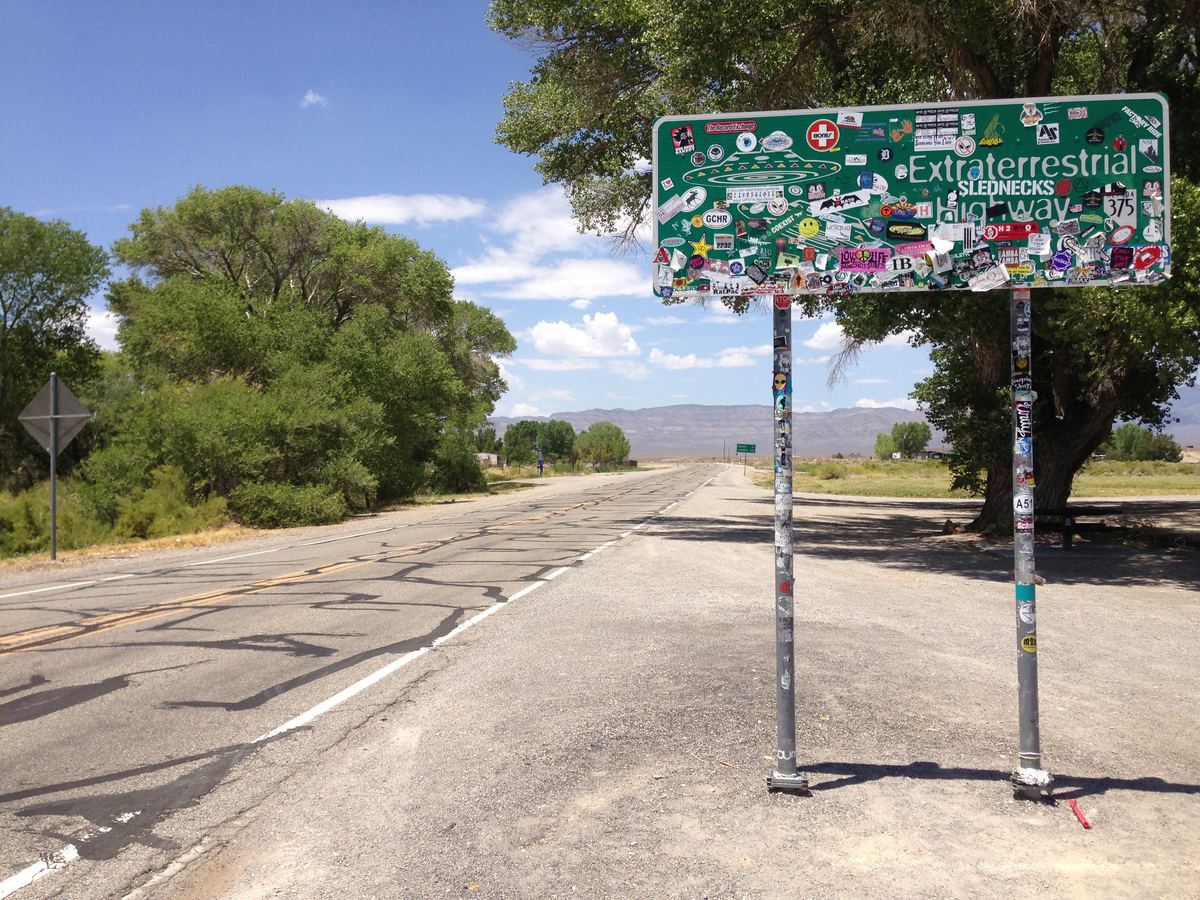 Sticker-covered green Extraterrestrial Highway sign beside a desert road with trees and distant mountains
