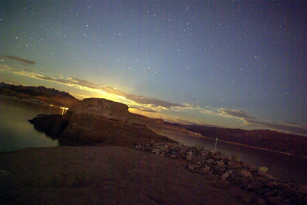 Starry night sky over rocky desert shoreline and calm water, with moonlight glowing behind distant mountains