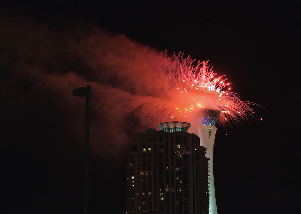A high-rise residential tower stands beside a tall observation tower at night as red fireworks burst overhead