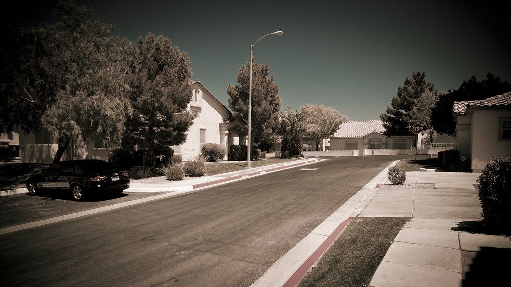 A quiet residential street lined with stucco houses, trees, a streetlight and a parked car on a sunny day