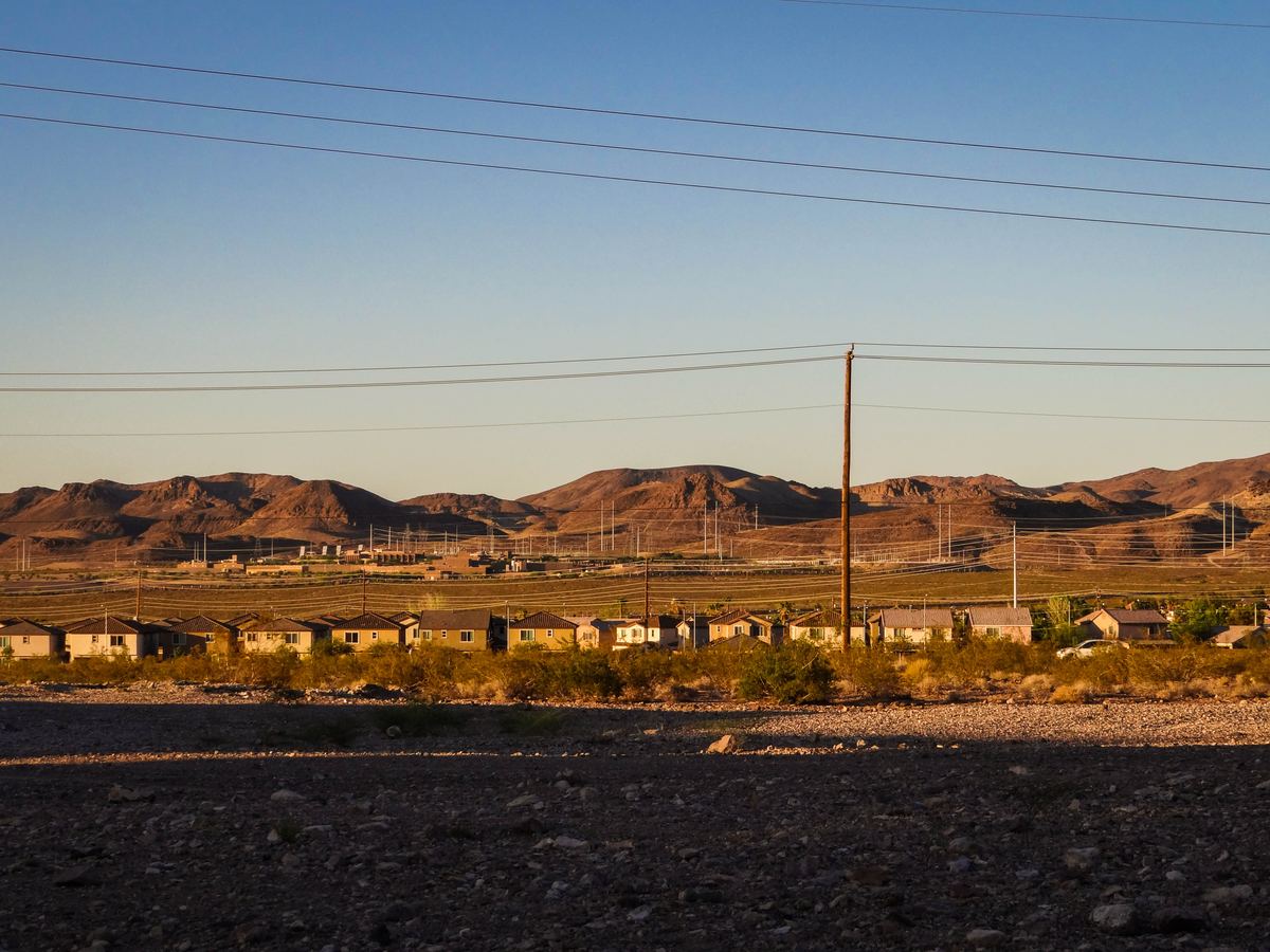 Row of desert homes beneath power lines with rocky open ground in front and brown mountains behind at sunset