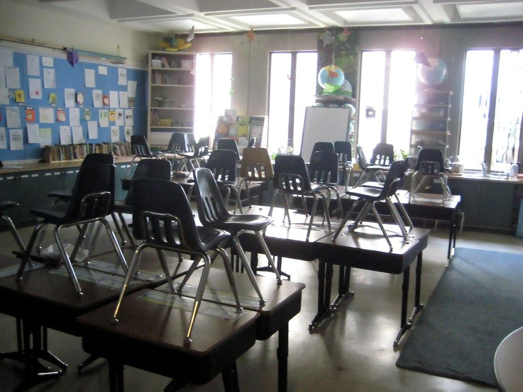 Empty classroom with chairs upturned on desks, a bulletin board, bookshelves and bright windows