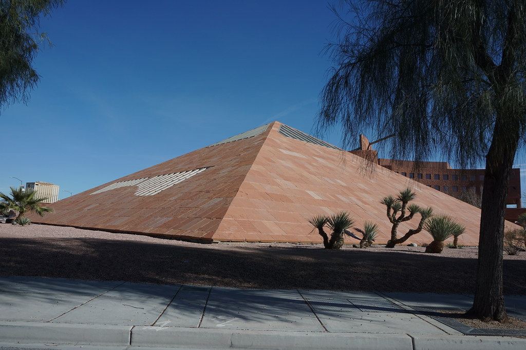 A sandstone pyramid structure beside a red office building, with desert plants, trees and a sidewalk under blue sky