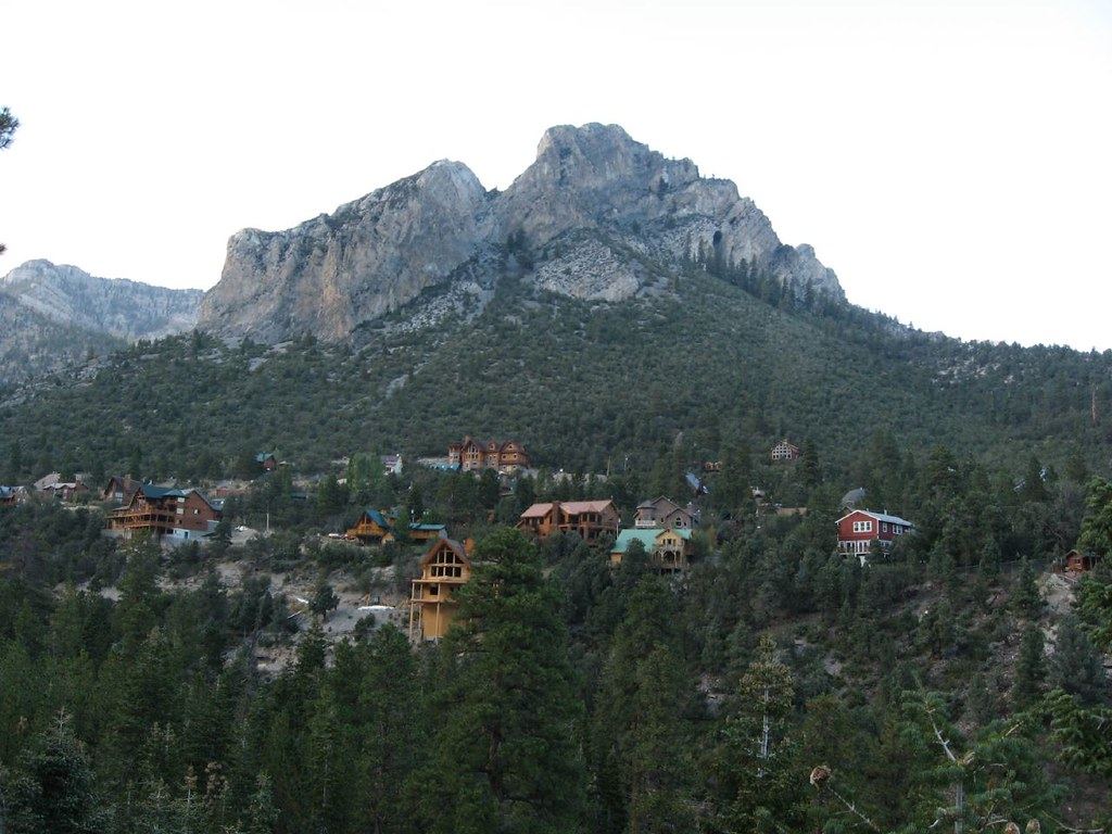 Wooden houses and cabins dot a pine-covered slope below a rugged gray mountain peak