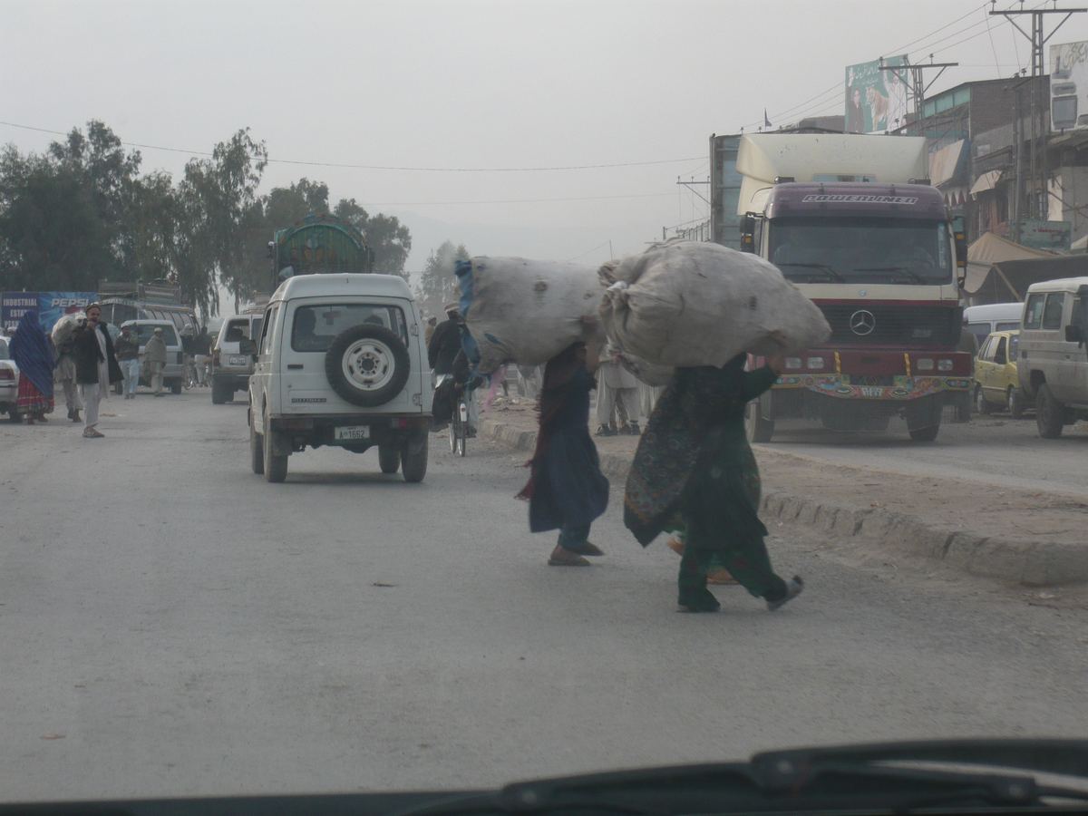 A busy street with trucks and a white SUV as two people carry large sacks on their heads across the road