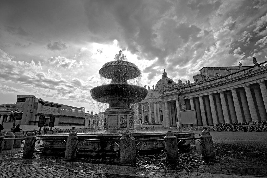 Black-and-white view of a fountain in St. Peter's Square with the colonnade and basilica dome under dramatic clouds