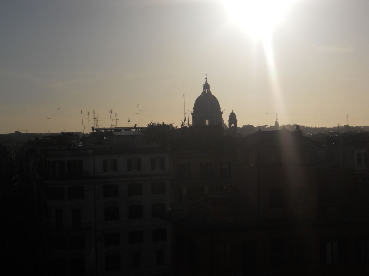Silhouetted Rome rooftops and a church dome against a hazy sky with bright sun and birds overhead