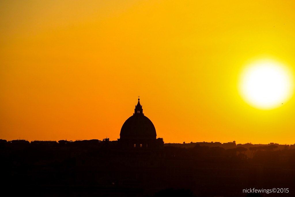 Silhouette of a large church dome against an orange sky with a bright sun setting over a city skyline