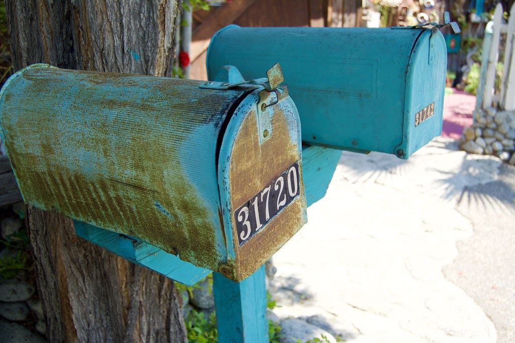 Two weathered turquoise mailboxes with house numbers mounted on a post beside a tree