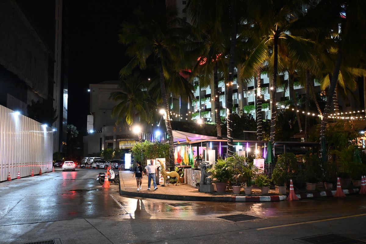A wet city street at night beside palm trees, string lights and an outdoor seating area, with two people walking
