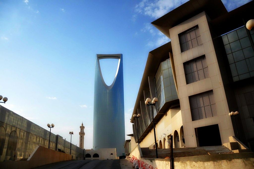 The Kingdom Centre tower in Riyadh rises behind modern buildings and a minaret under a blue sky