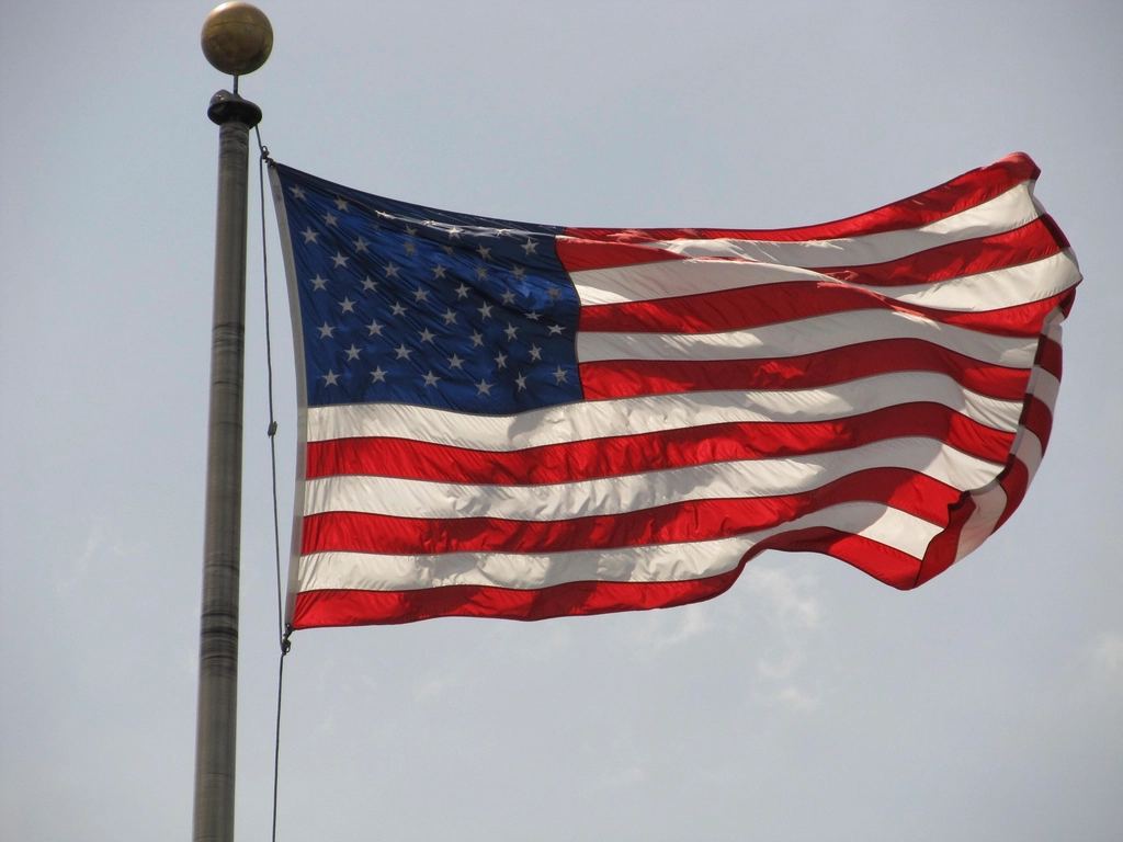 An American flag waves from a metal flagpole against a pale, lightly clouded sky