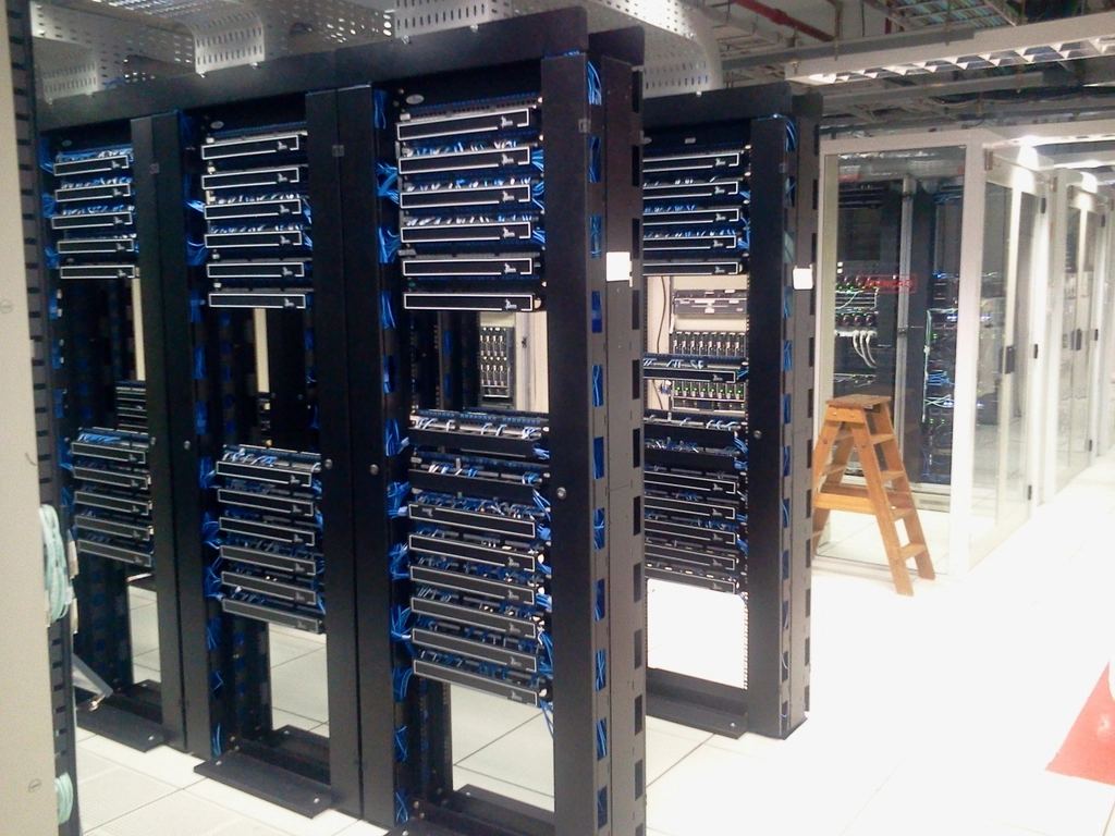 Rows of black network racks with blue cables in a server room, with a wooden stepladder near glass enclosures