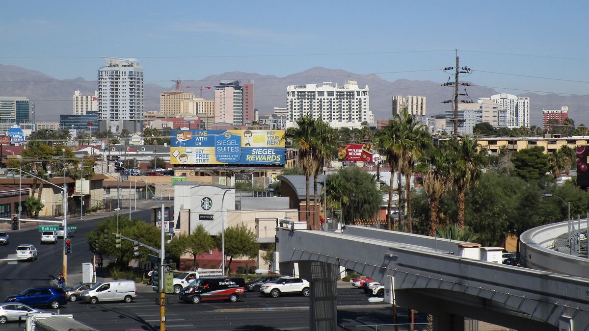 Las Vegas street intersection with palm trees, an elevated monorail track, and downtown high-rises against mountains