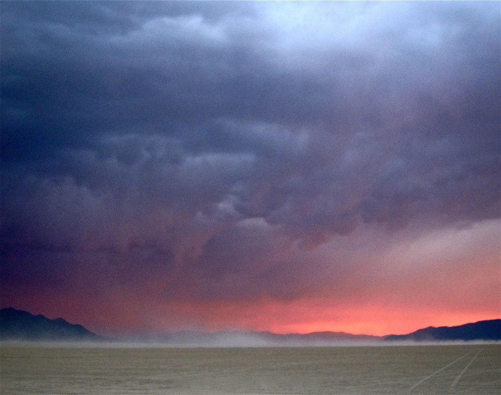 Dark storm clouds over a flat desert plain with mountains and a pink glow on the horizon