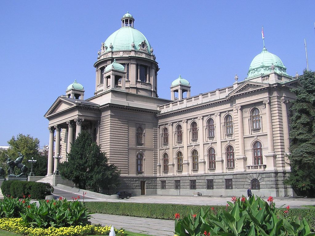 A large domed neoclassical parliament building with green copper roofs behind flowerbeds under a clear blue sky