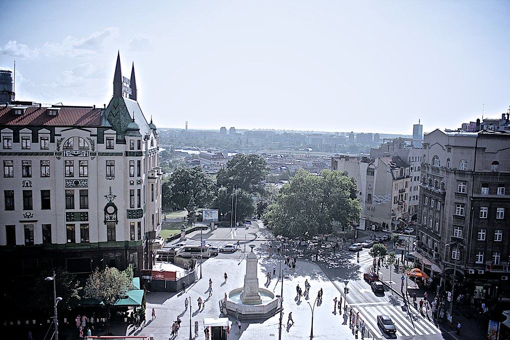 Elevated view of a city square with a fountain, an ornate corner hotel, pedestrians and a distant skyline