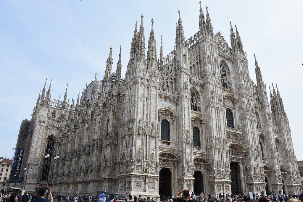 The ornate white Gothic facade and spires of Milan Cathedral, with people gathered in the square below