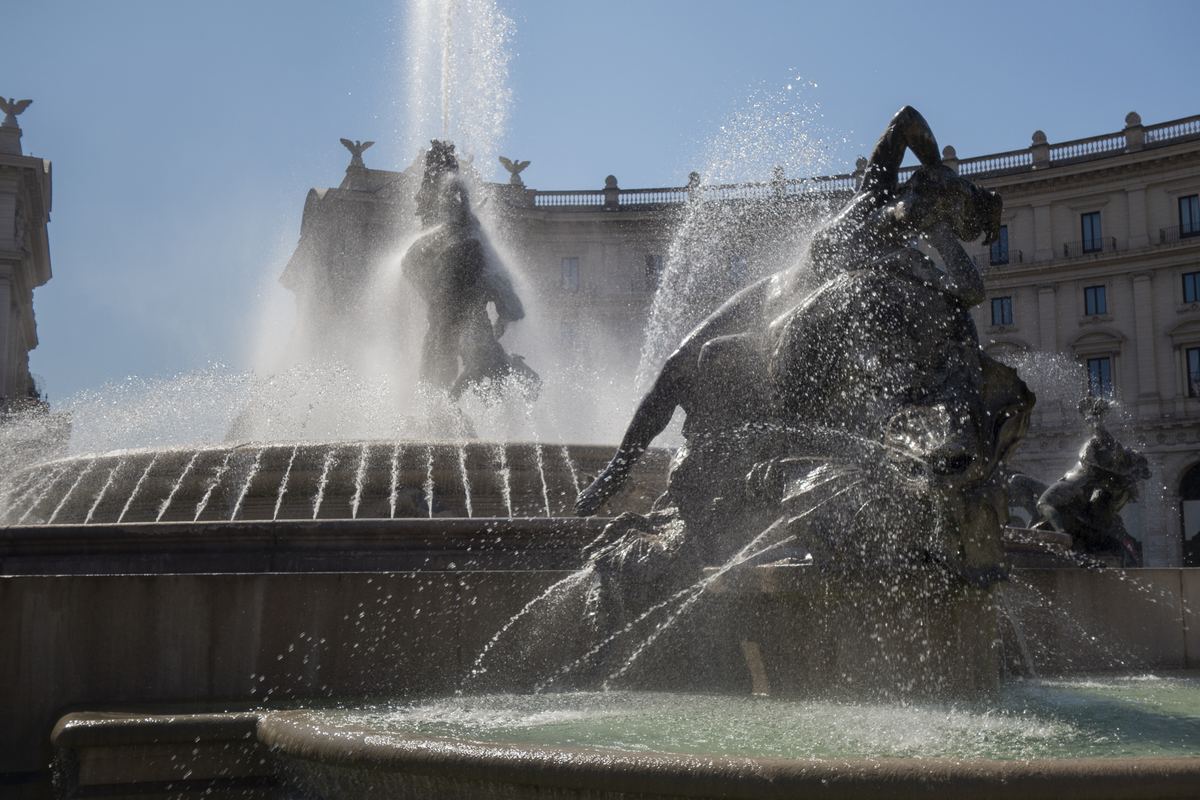Bronze sculptures and spraying water at a fountain in front of a curved neoclassical building under a blue sky