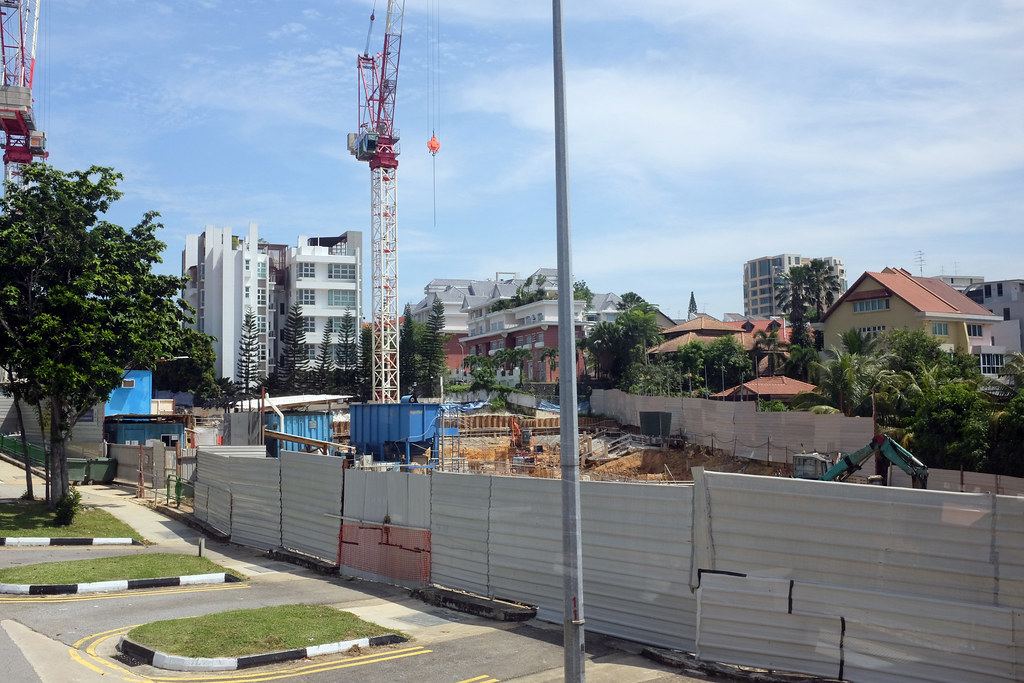 Construction site behind metal hoarding, with a tower crane, an excavator and apartment buildings under a blue sky