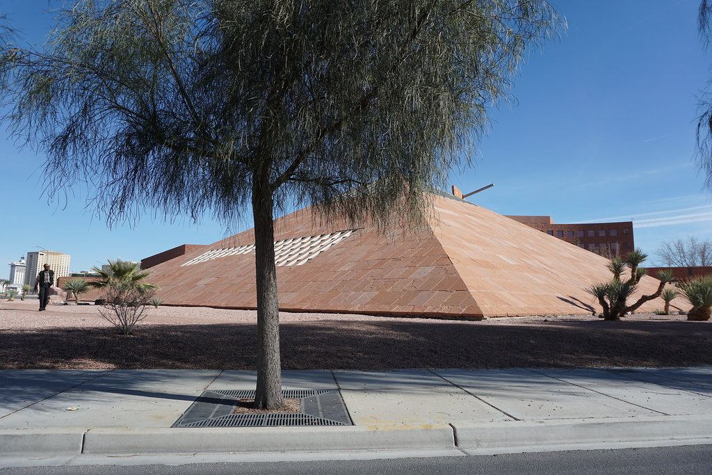 A red sandstone pyramid-shaped building with desert landscaping behind a tree-lined sidewalk under a blue sky