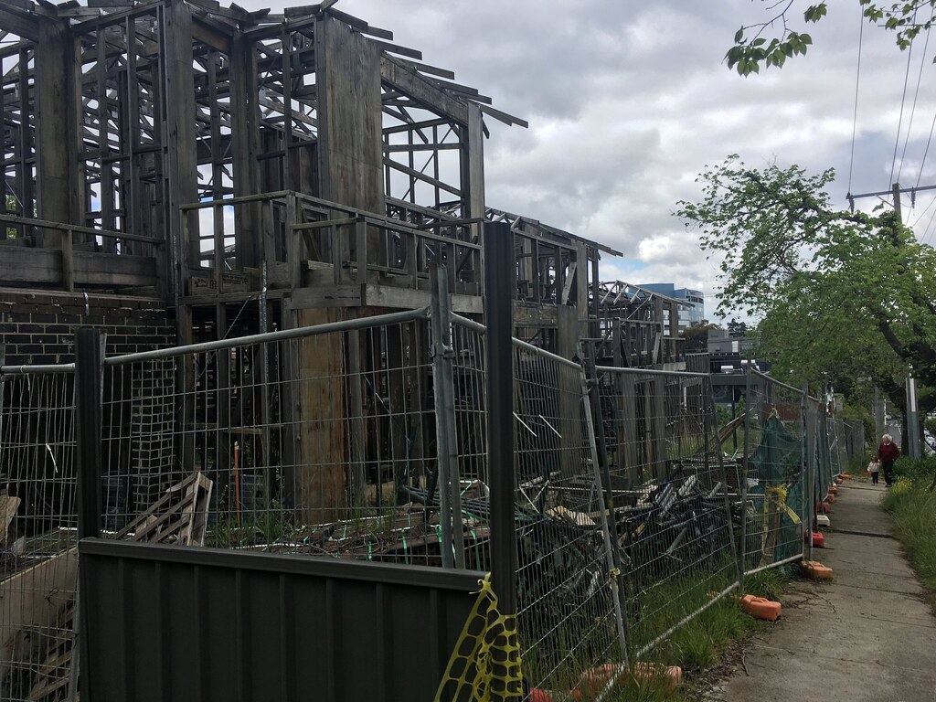 Weathered timber frames of unfinished townhouses behind temporary metal fencing beside a sidewalk