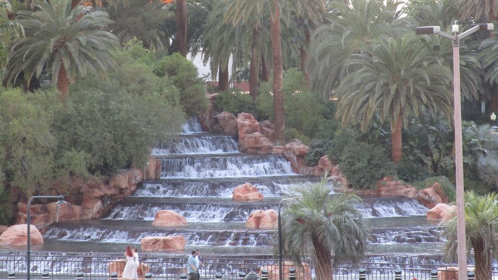 Tiered waterfalls cascade over red rocks beneath tall palm trees, with a few people walking along a railing