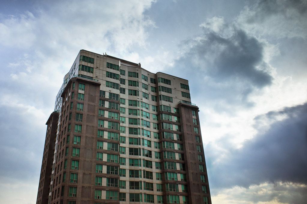 A tall brown and beige residential tower with rows of green-tinted windows against a cloudy sky