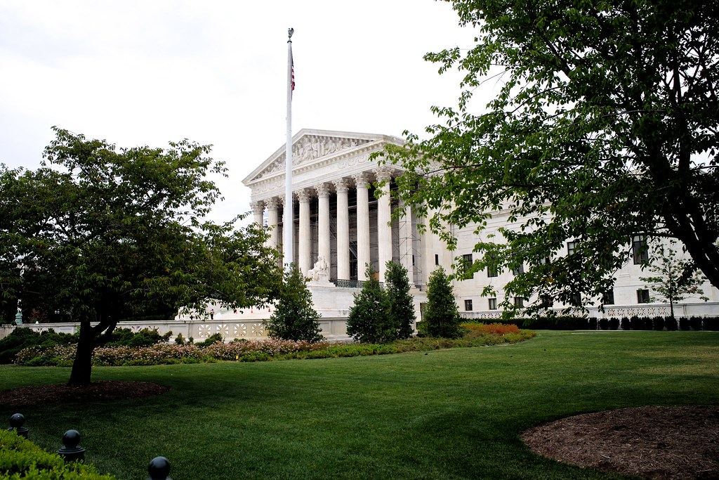 The white-columned US Supreme Court building behind a flagpole, trees and a green lawn on a cloudy day