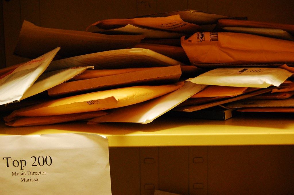 A messy pile of padded mailers and envelopes on a yellow shelf, with a paper sign taped below