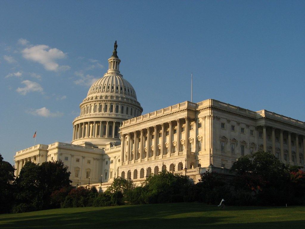 The U.S. Capitol dome and Senate wing in warm sunlight above a green lawn and trees under a blue sky