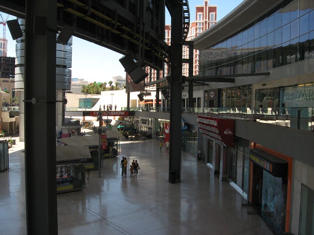 Wide view of an open-air shopping mall plaza with storefronts, kiosks and a few shoppers under a steel canopy