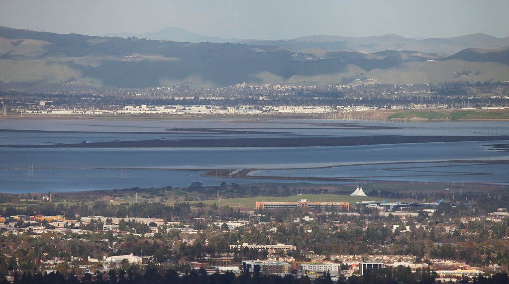 Elevated view of suburban buildings and offices beside San Francisco Bay salt flats, with hills in the distance