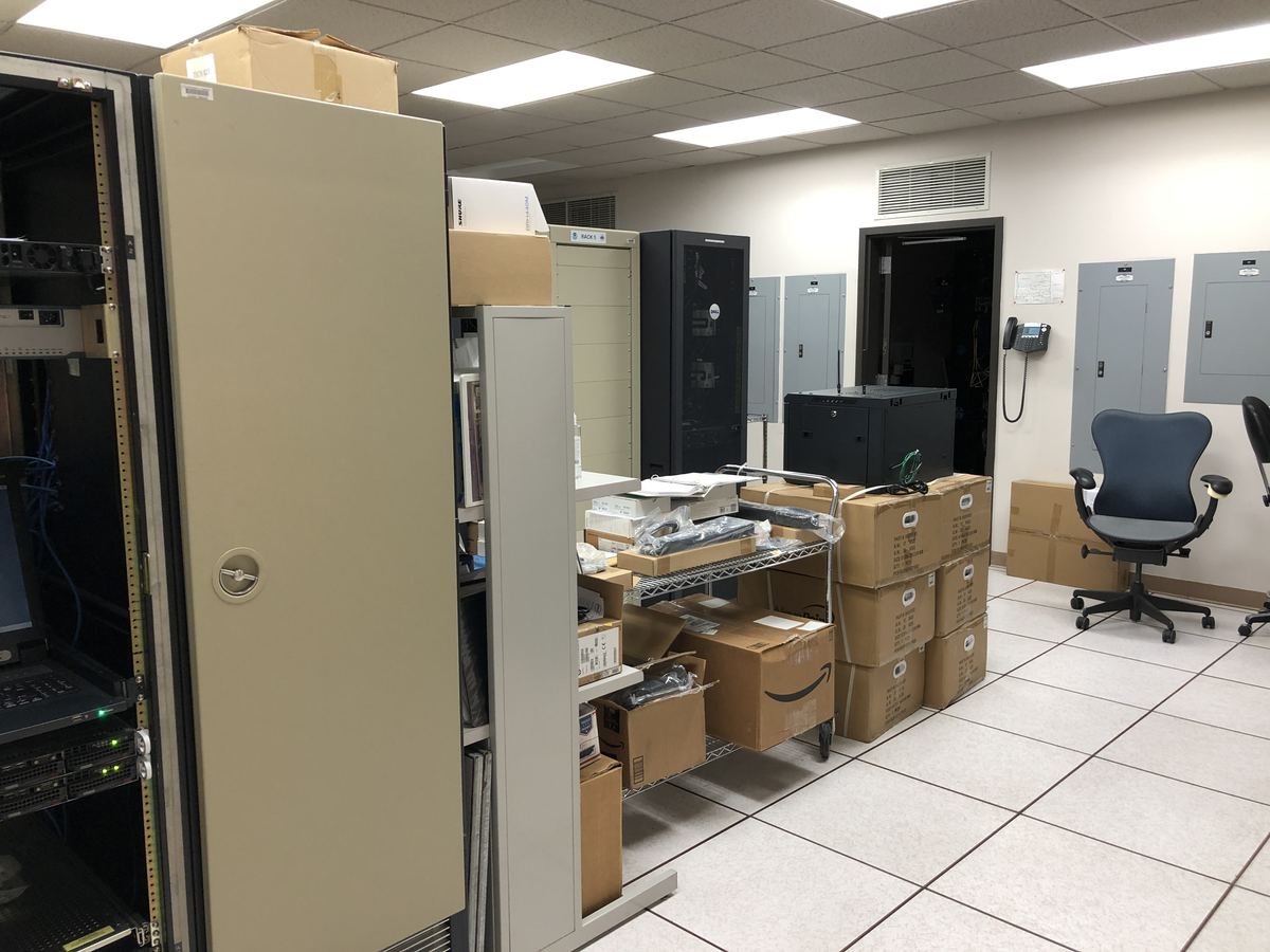 Server racks, stacked cardboard boxes and an office chair in a white-tiled computer equipment room