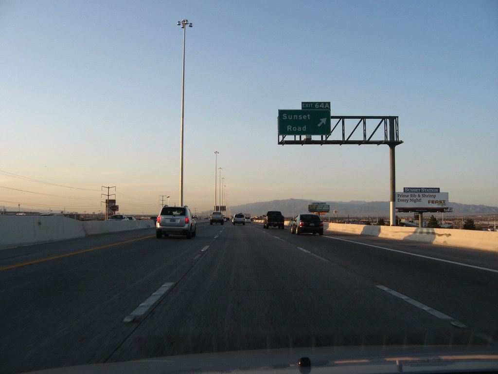 Cars travel a multi-lane highway at dusk beneath a green Sunset Road exit sign, with desert mountains ahead