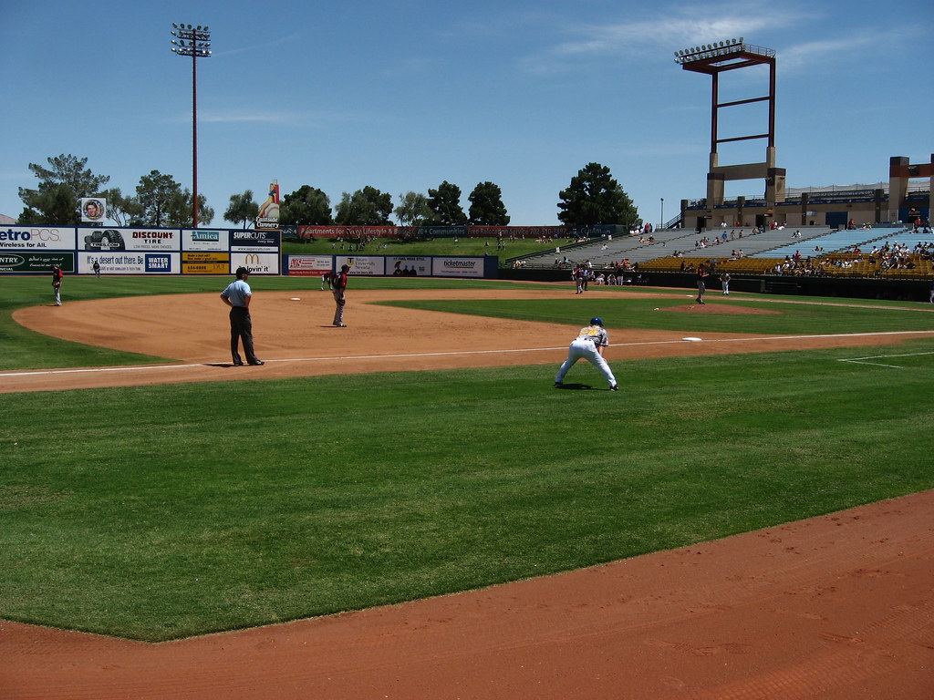 Baseball game under way on a sunny day at a minor league ballpark with grandstands and light towers
