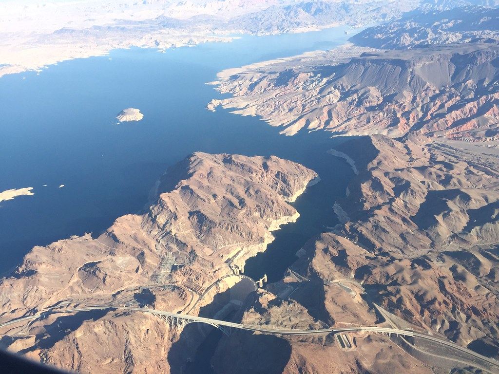 Aerial view of a desert lake narrowing into a canyon, with a dam and an arched highway bridge below