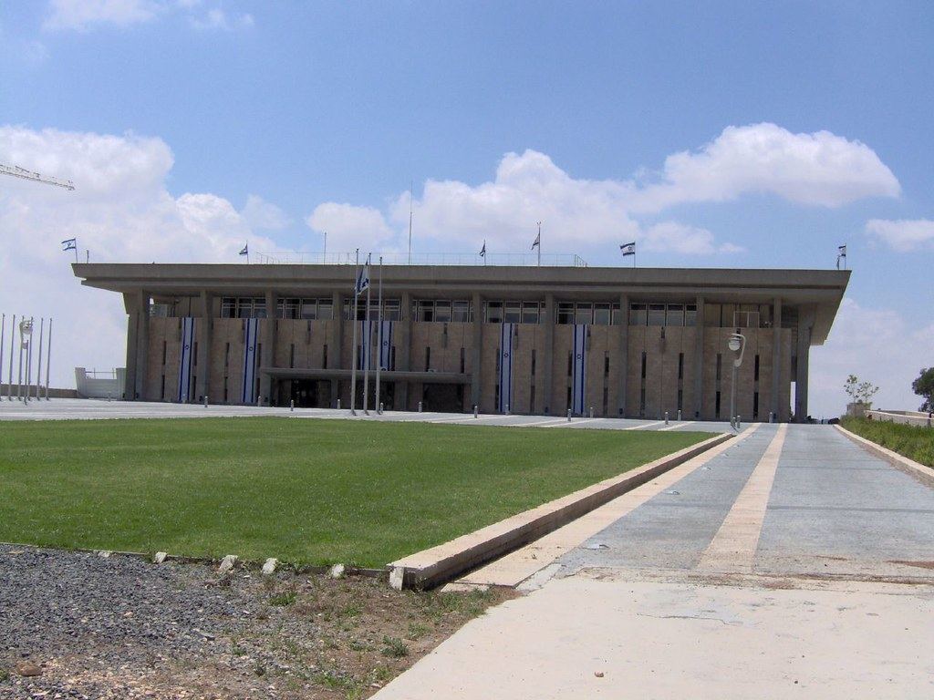 A large flat-roofed stone building with Israeli flags and banners, a lawn and a paved walkway in front
